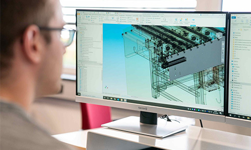 Person mit Brille vor zwei Monitoren, auf einem Bildschirm eine technische CAD-Zeichnung eines komplexen mechanischen Bauteils