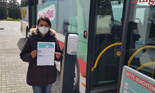 Person mit winterlicher Jacke und Mund-Nasen-Schutz, die ein Dokument in der Hand hält, steht vor einem Testbus und einem Desinfektionsspender auf einem gepflasterten Bereich