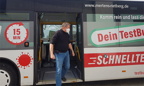 Mann mit Schutzmaske steigt aus einem Bus mit Aufschrift zum Corona-Schnelltest und Hinweis auf 15 Minuten Wartezeit.