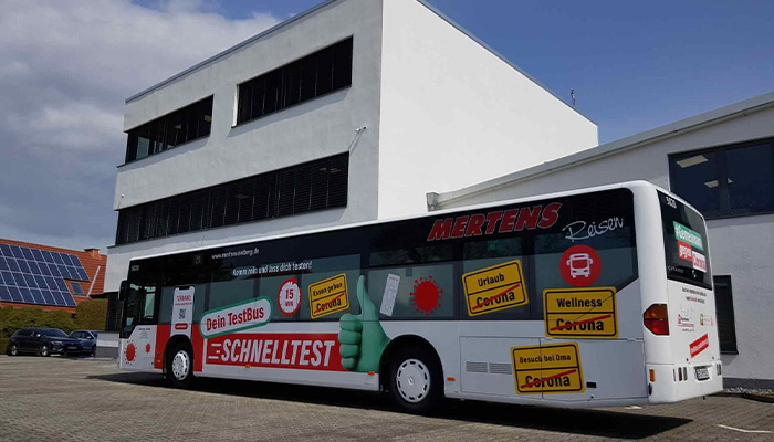 Großer, weißer Reisebus mit Werbeaufschrift für Schnelltests und diversen gelben Corona-Warnhinweisschildern, geparkt vor einem weißen Bürogebäude an einem sonnigen Tag.