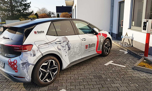 Weißer Elektro-SUV mit Grafiken und dem Logo von Rotte, der vor einem modernen Gebäude an einer Ladestation für Elektrofahrzeuge steht.
