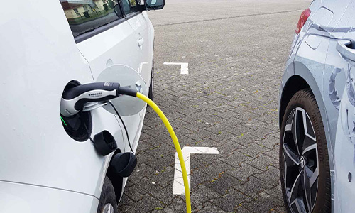 Zwei geparkte Elektroautos, eines von links mit gelbem Ladekabel an der Ladestation verbunden, sichtbare Pflastersteine am Boden und weiß markierte Parklinien