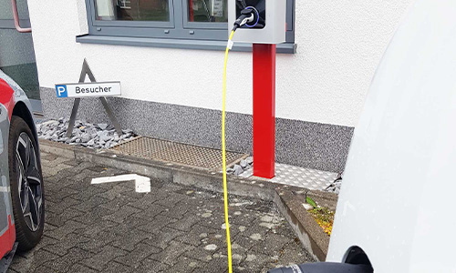 Elektroauto lädt an einer Ladestation, Kabel gelb, sichtbar sind Teile zweier Fahrzeuge, im Hintergrund ein Wegweiser mit dem Wort 