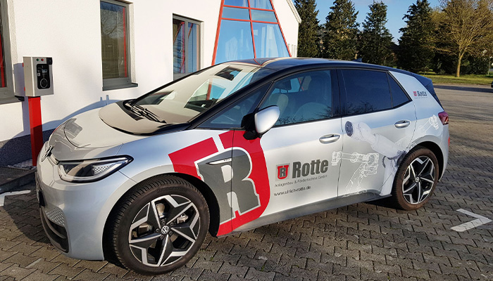 Silbernes Elektroauto von Volkswagen mit Werbeaufdrucken der Firma Rotte, geparkt auf gepflastertem Parkplatz neben einem Gebäude mit weißen Wänden und roten Fensterrahmen.