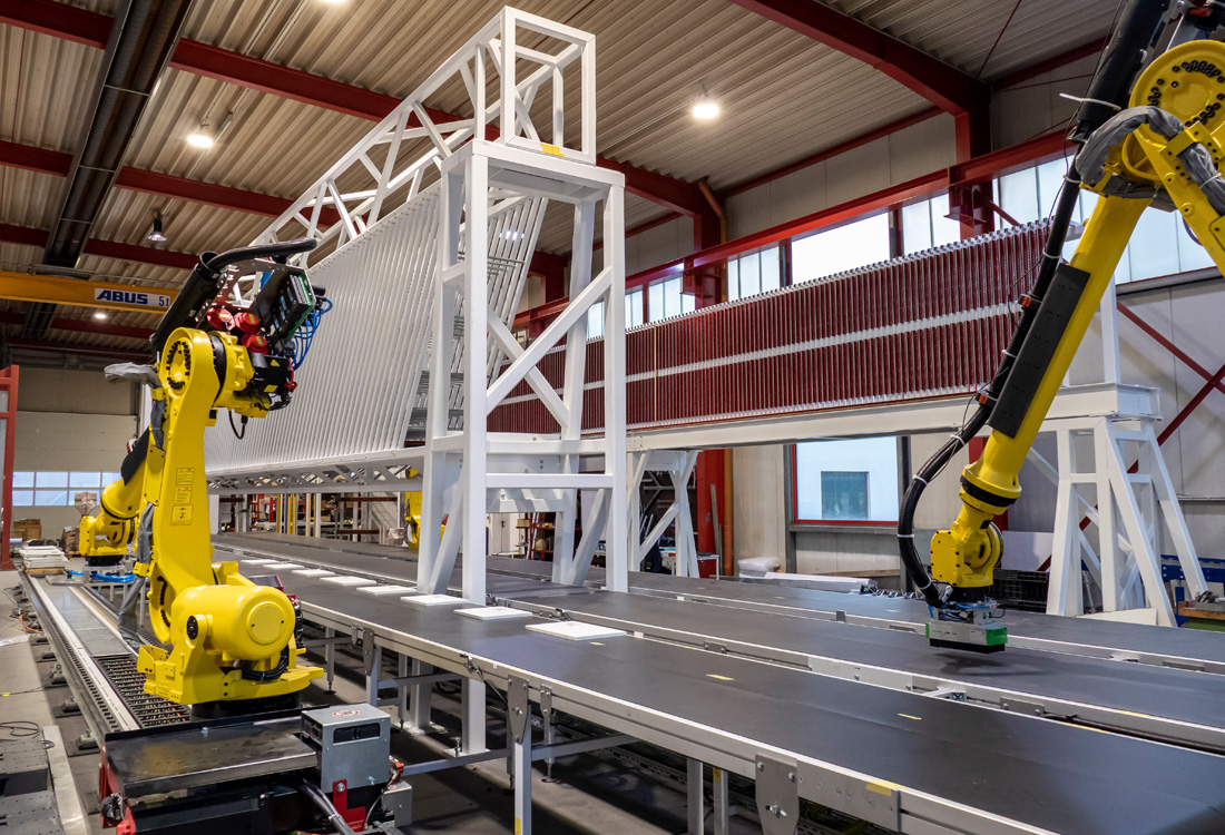 Industrielle Fertigungshalle mit zwei gelben Industrierobotern, die an einem langen Förderband mit weißen Stäben und einer weißen Stahlstruktur arbeiten.