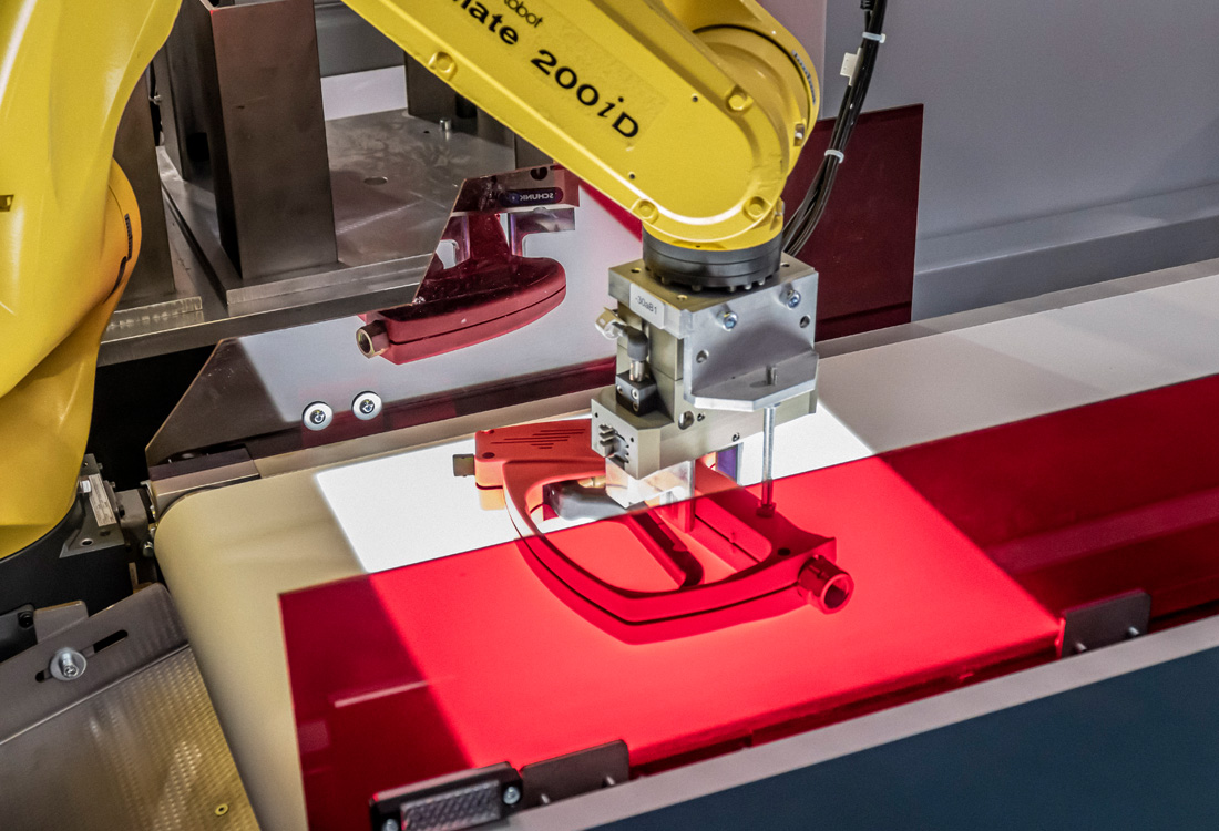 Gelber Industrieroboterarm mit Aufschrift "mate 200iD" bearbeitet ein rotes Kunststoffteil auf einem Förderband unter roter Beleuchtung in einer Produktionsumgebung.