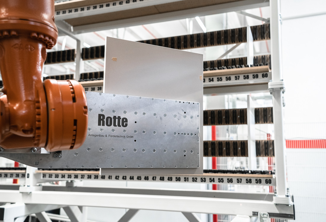 Industrieroboterarm hält Metallplatte mit Aufschrift 'Rotte Anlagenbau & Fördertechnik GmbH' vor einem nummerierten Regal in einer Fabrikumgebung