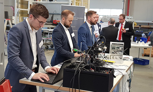 Fünf Männer in Anzügen arbeiten an einem Tisch mit technischen Geräten und Laptops in einer Halle, zwei Männer im Hintergrund unterhalten sich stehend.
