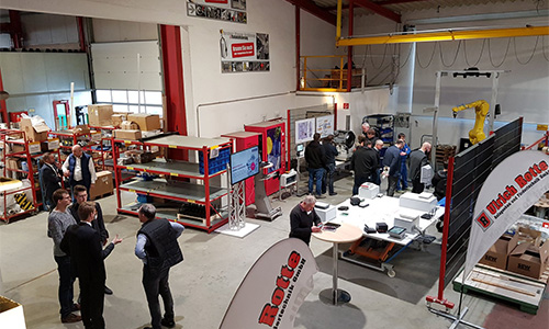 Blick in eine Industriehalle mit mehreren Personen in Gesprächen, Regalen, einem Roboterarm und Tischen mit technischen Geräten, dazu Werbebanner der Firma Rotte
