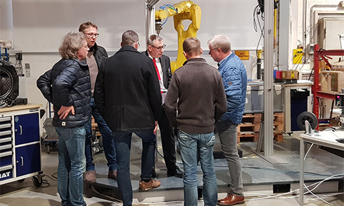 Sechs Männer stehen in einer Industriehalle um eine Maschine oder ein Gerät herum und diskutieren, im Hintergrund ein gelber Roboterarm und Regale mit Werkstattmaterialien.
