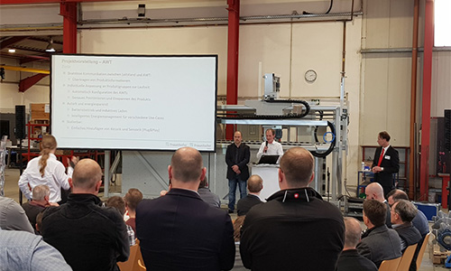 Menschen in einem Konferenzraum bei einer Präsentation mit Projektorbildschirm und Rednern, technische Ausrüstung und Industrielampen im Hintergrund