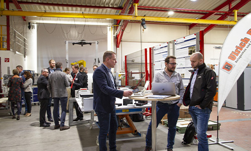 Menschen in einem industriellen Ausstellungs- oder Messestand mit technischen Displays und Fahne, einzelne Gruppen unterhalten sich stehend in lockerer Atmosphäre.