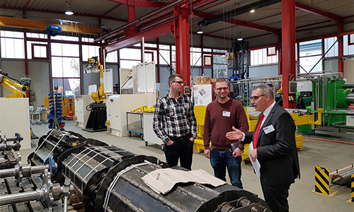 Drei Männer in einer Industriehalle stehen neben einer großen Maschinenkomponente, umgeben von technischen Anlagen und Ausrüstung.