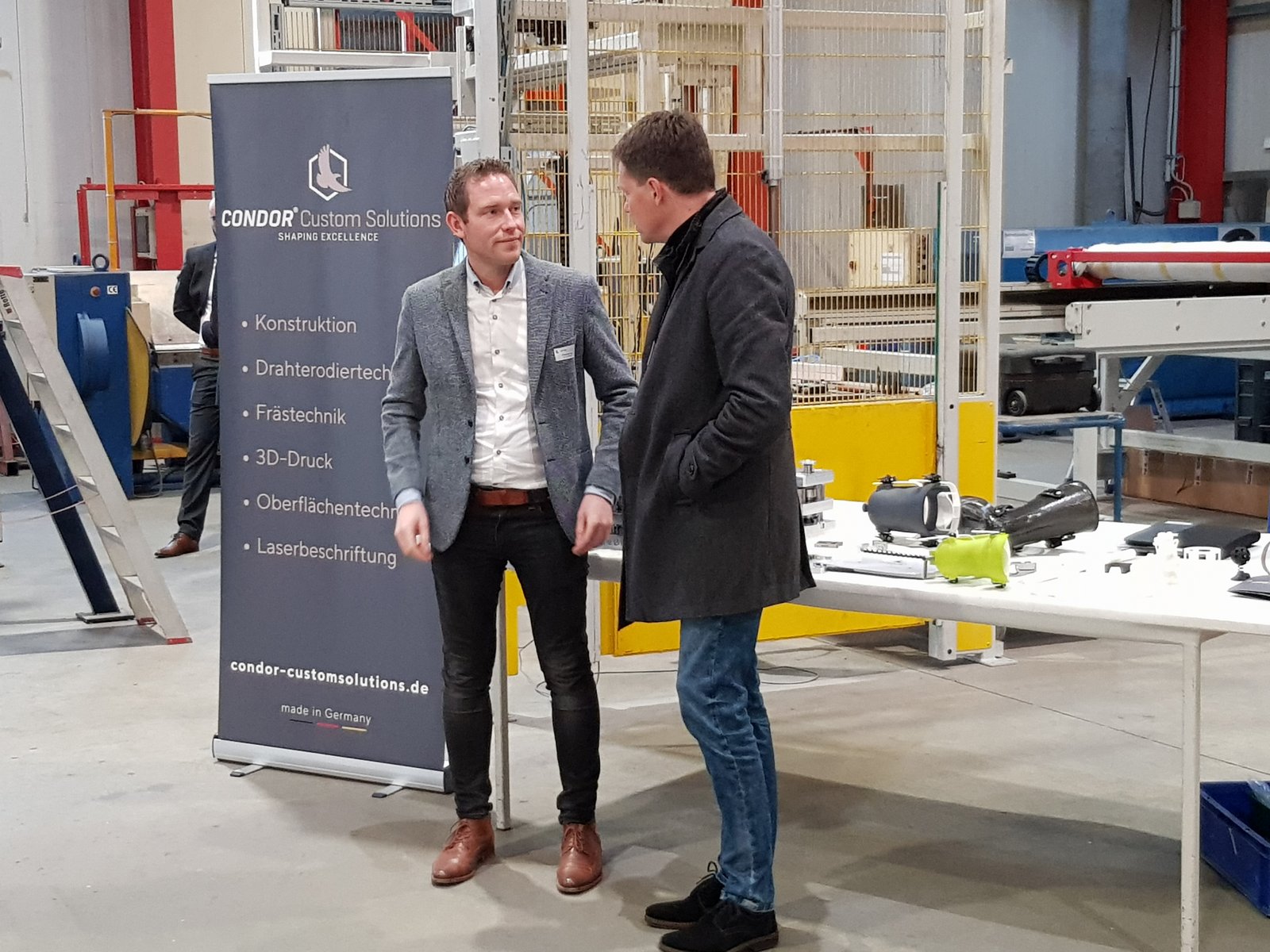Zwei Männer im Gespräch vor einem Banner der Firma Condor Custom Solutions in einer Produktionshalle mit Industriemaschinen und einem Tisch mit Bauteilen.