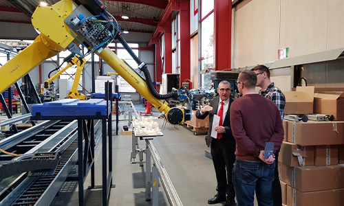 Eine Fabrikumgebung mit einem großen gelben Industrieroboterarm und drei Personen, die auf den Roboterarm blicken und diskutieren, umgeben von Kartons und Maschinen.