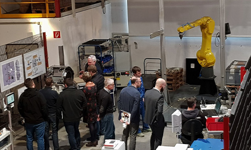 Gruppe von Menschen, vorwiegend Männern, in einer Werkstatt oder Fabrikumgebung mit Maschinen und einem gelben Industrieroboterarm im Hintergrund.