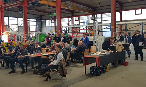 Gruppe von Menschen, die in einer großen Werkhalle auf Stühlen und vor Tischen sitzen und stehen, bei einer Veranstaltung oder Präsentation mit Getränken und Snacks auf einem Tisch in der Nähe.