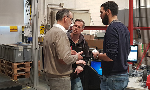 Drei Männer in einer Werkstatt oder einem Labor im Gespräch, umgeben von technischen Geräten und Regalen mit Kisten auf Paletten.