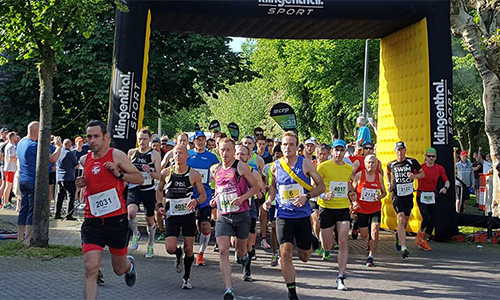 Läufer unterschiedlicher Altersgruppen in Sportkleidung starten bei einem Laufwettbewerb unter einem schwarzen und gelb markierten Startbogen mit der Aufschrift Klingenthal SPORT, umgeben von Bäumen und Zuschauern am Straßenrand.