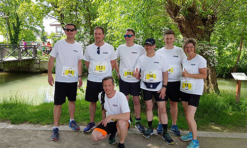 Eine Gruppe von sechs Personen in weißen T-Shirts und schwarzen Shorts mit Laufnummern posiert lächelnd im Freien vor einem kleinen Fluss und einer Brücke, umgeben von Bäumen und Grünflächen.