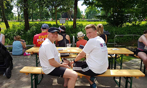 Zwei Männer in Sportkleidung sitzen an einem Holztisch im Freien, umgeben von weiteren Personen auf Bänken, mit grüner Parklandschaft im Hintergrund.