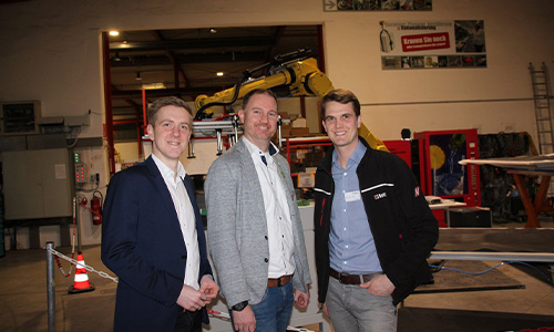 Drei Männer in einem Industrie- oder Laborumfeld vor einem großen Roboterarm, zwei davon tragen Blazer, der mittlere trägt einen grauen Cardigan.