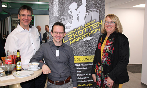 Drei lächelnde Personen stehen vor einem Banner mit der Aufschrift LZKOTTEN MARATHON, links auf einem Stehtisch stehen Getränke und eine Schüssel.