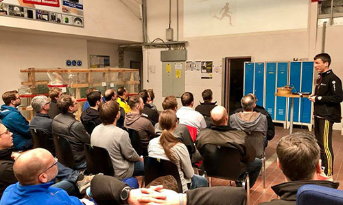 Gruppe von Menschen in einem Raum, die einem stehenden Mann zuhören, der vor blauen Schließfächern spricht, mit technischer Ausrüstung im Hintergrund.