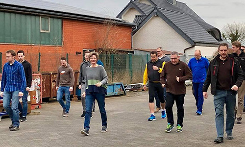 Gruppe von etwa zwölf Personen unterschiedlichen Alters, die draußen auf einem Gehweg in einem Wohngebiet spazieren gehen, mit Gebäuden im Hintergrund.