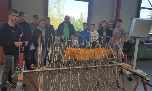 Gruppe von Jugendlichen und einem Erwachsenen in einer Werkstatt, die an einer Metallkonstruktion mit mehreren vertikalen und gebogenen Stäben sowie einem orangefarbenen Element arbeitet, im Hintergrund Fenster mit Blick auf Bäume.