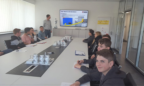Junge Menschen sitzen an einem langen weißen Konferenztisch in einem modernen Besprechungsraum, ein Mann steht vor einem Bildschirm mit einer Präsentation und einem Pokal neben dem Bildschirm.