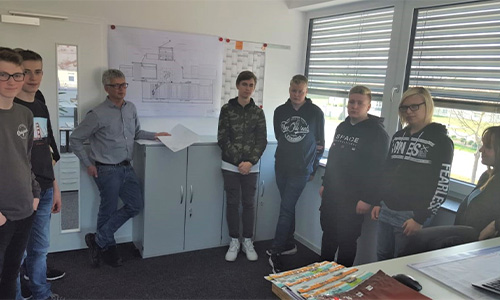 Sieben Personen, einschließlich eines älteren Mannes und sechs jüngeren Menschen, die in einem Raum mit Schrank und großen Fenstern stehen, an der Wand Zeichnungen und Pläne hängen.
