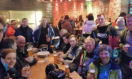 Menschen sitzen und stehen um einen langen Holztisch in einer gemütlichen Gaststätte, viele halten Biergläser, im Hintergrund Holzscheiben an der Wand und eine Tafel mit Beschriftungen