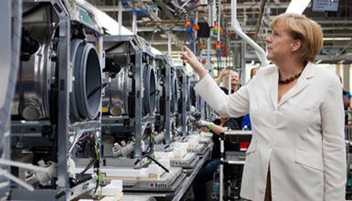 Frau im weißen Blazer zeigt auf eine Reihe von Waschmaschinen in einer Fabrikhalle, während andere Arbeiter im Hintergrund tätig sind.