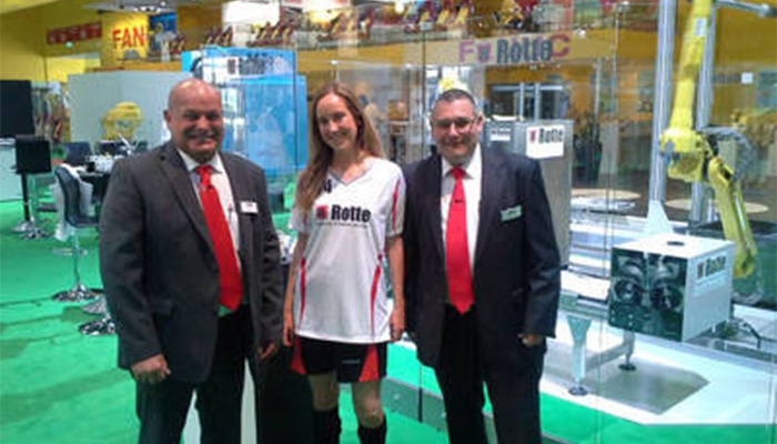 Drei Personen, zwei Männer in Anzügen mit roten Krawatten und eine Frau in sportlichem Outfit mit Firmenlogo, stehen vor einem Messestand mit automatisierter Robotertechnik und grüner Bodenfläche.