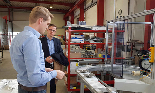 Zwei Männer in einem Lager oder einer Werkstatt betrachten eine technische Anlage mit einem Förderband und einer transparenten Schutzvorrichtung in einer industriellen Umgebung.