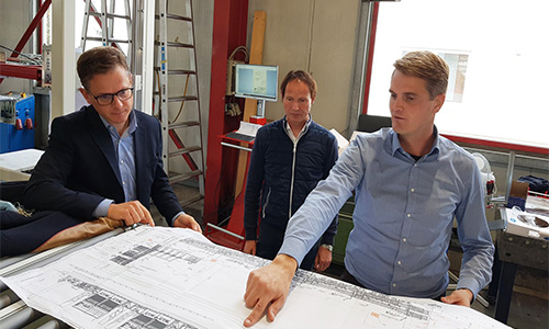 Drei Männer in einem Arbeitsraum schauen gemeinsam auf einen großen technischen Plan, einer zeigt mit dem Finger darauf.