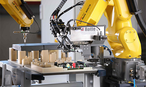 Gelbe Industrieroboterarme montieren oder bearbeiten elektronische Baugruppen auf einer Werkbank in einer Fabrikumgebung.