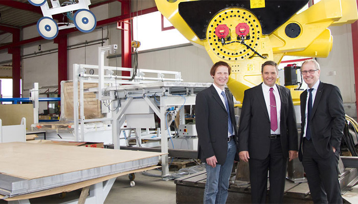 Drei Männer in Anzügen stehen in einer Industriehalle vor einer großen, gelben Maschine mit Kabeln, umgeben von weiteren technischen Geräten und Werkbänken.