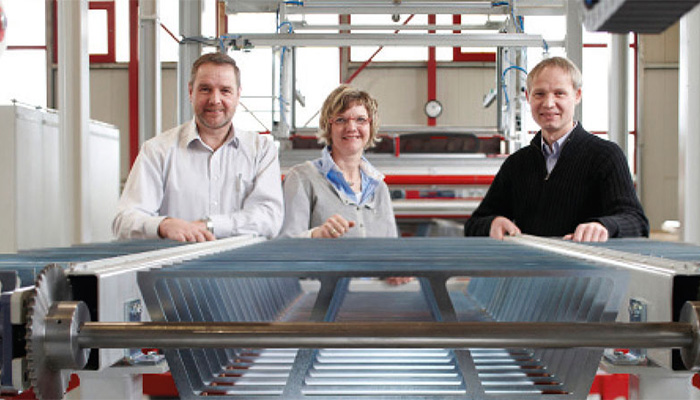Drei Personen stehen nebeneinander in einer industriellen Umgebung mit Metallstruktur im Vordergrund, sie lächeln und blicken in die Kamera.