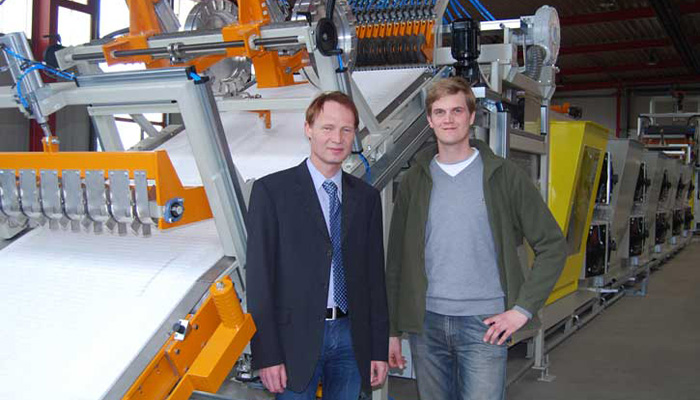 Zwei Männer, einer im Anzug und einer mit grüner Jacke, stehen in einer Industriehalle vor einer großen, komplexen Maschine mit orangefarbenen und gelben Elementen.
