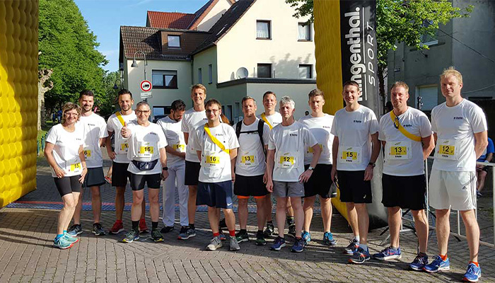 Gruppe von 14 Menschen in Sportkleidung mit Startnummern, die vor einem aufblasbaren Tor mit Markenlogo für ein Sport-Event posieren, im Hintergrund Häuser und Bäume an einem sonnigen Tag.