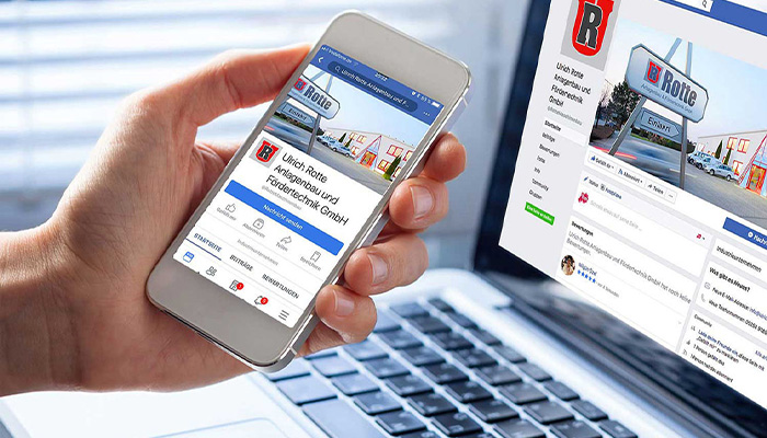 Hand hält ein Smartphone, auf dem die Facebook-Seite von Ulrich Rotte Anlagenbau und Fördertechnik GmbH angezeigt wird, im Hintergrund ist ein geöffneter Laptop mit derselben Facebook-Seite sichtbar