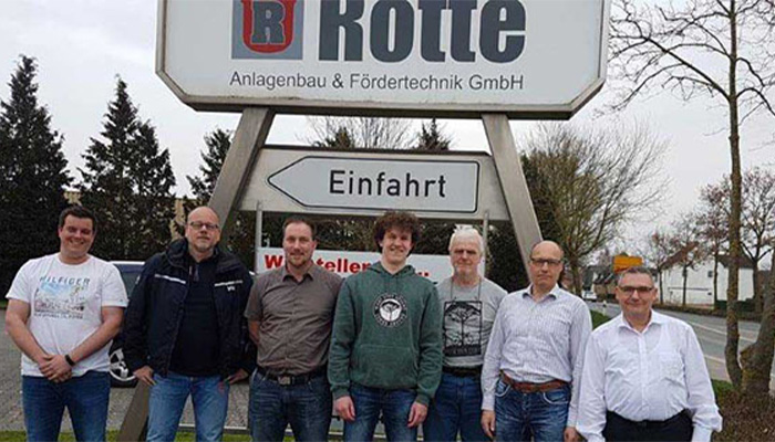 Sieben Männer, die vor einem großen Firmenschild mit der Aufschrift 'Rolle Anlagenbau & Fördertechnik GmbH' und einem Pfeilschild mit der Aufschrift 'Einfahrt' stehen, im Hintergrund Bäume und ein Parkplatz.