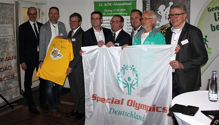 Acht Männer stehen in einem Innenraum, einige halten ein Banner mit der Aufschrift "Special Olympics Deutschland", einer zeigt ein gelbes Trikot, im Hintergrund sind Plakate mit Veranstaltungshinweisen zu sehen.