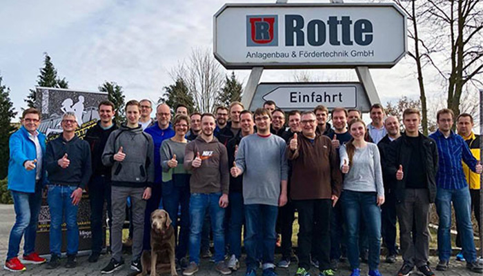 Gruppenfoto von etwa 24 Personen und einem Hund, die vor einem Firmenschild von Rotte Anlagenbau & Fördertechnik GmbH stehen und Daumen nach oben zeigen.
