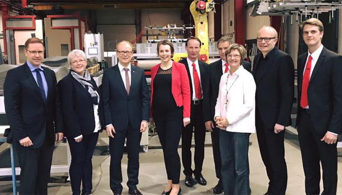 Neun Personen in formeller Kleidung stehen in einer Industriehalle vor Maschinen und schauen in die Kamera.
