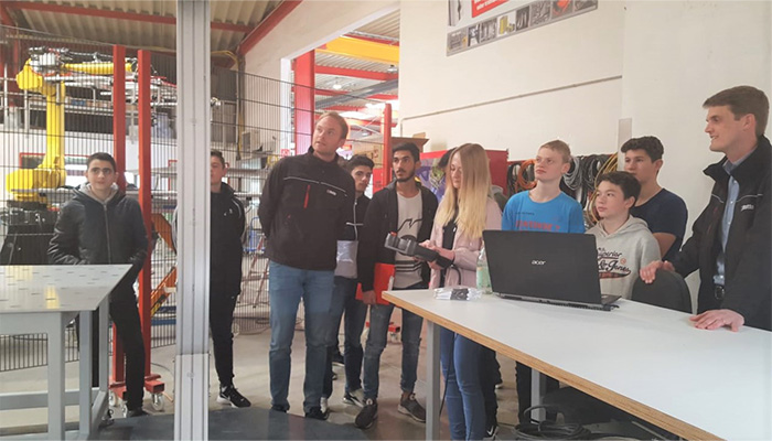 Gruppe von jungen Erwachsenen und Jugendlichen in einer Industriehalle, einige stehen neben einem Tisch mit einem Laptop, eine Person hält ein Kameraobjektiv, andere blicken interessiert in eine Richtung.