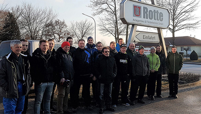 Gruppe von 16 Personen, überwiegend Männer, vor einem Firmenschild mit der Aufschrift »Rotte Anlagenbau & Fördertechnik GmbH« und mehreren Schildern, stehend auf einem Gehweg an einem sonnigen Wintertag mit kahlen Bäumen im Hintergrund.