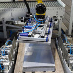Industrieroboter mit Greifmechanismus, der eine metallische Platte auf einem Förderband oder in einer Produktionslinie bewegt, umgeben von pneumatischen Komponenten und Sicherheitszaun.